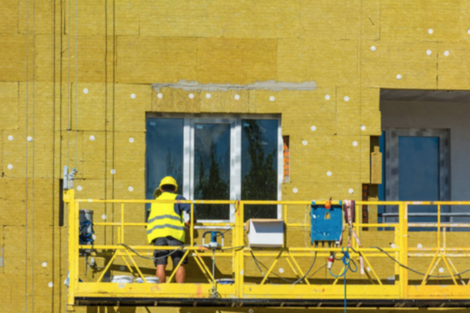 Worker with yellow hard hat wearing yellow vest on a yellow suspended platform installing insulation on the exterior of a high-rise building