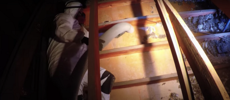 Worker in a white protective suit with respirator mask laying down in attic holding a white vacuum hose and removing flawed insulation