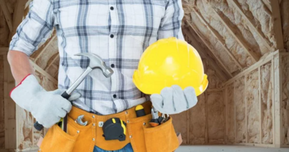 construction worker wearing gloves, holding a hammer in the right hand and a yellow helmet in the left hand, standing in house that is being insulated and renovated 