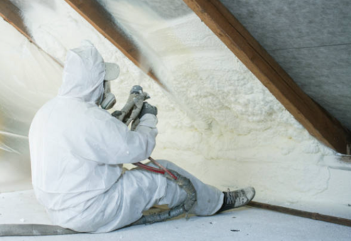 man in white protective suit wearing a respirator mask sitting while applying spray foam insulation to attic wall